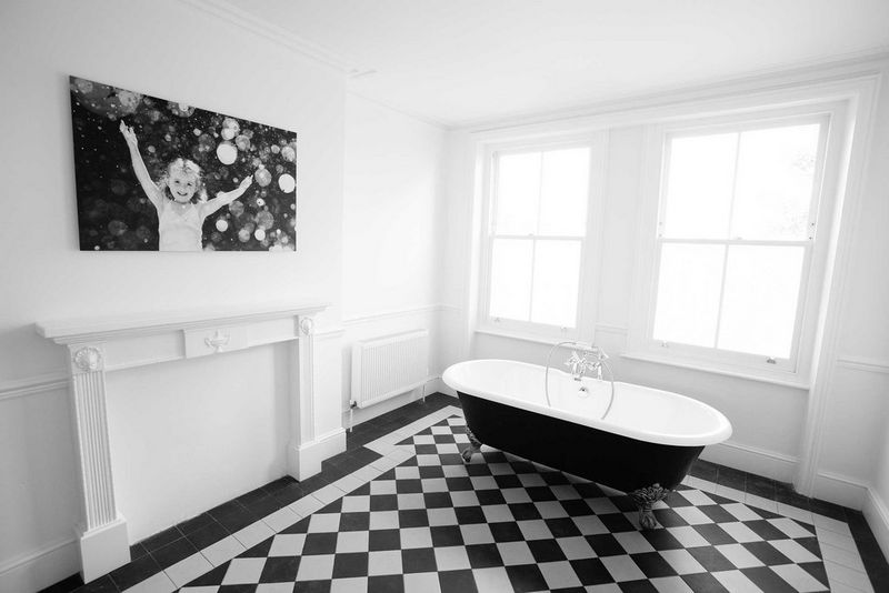 صورة مطبوعة على لوحة قماشية لفتاة تلعب بالفقاعات معروضة على جدار في حمام كبير جدرانه بيضاء وأرضيته مرسوم عليها مربعات.