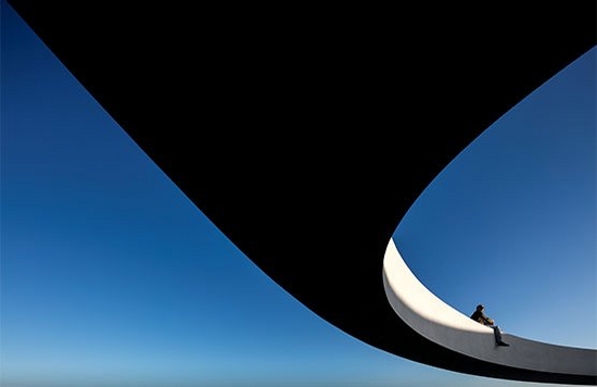 Against a bright blue sky, a man sits on the edge of a huge curving platform that's elevated several metres above the ground. 
