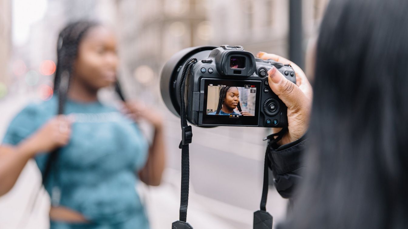امرأة بضفائر طويلة تلتقط صديقتها التي تحمل كاميرا EOS R6 من Canon صورًا لها في الشارع.
