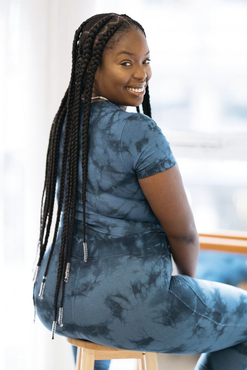 A woman with long braids sits facing away from the camera, with her head turned back towards it. She is wearing a matching tie-dye top and leggings.