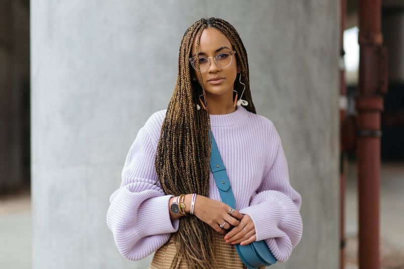 A woman with long braids and glasses wearing a lilac jumper and a small blue satchel across her body.