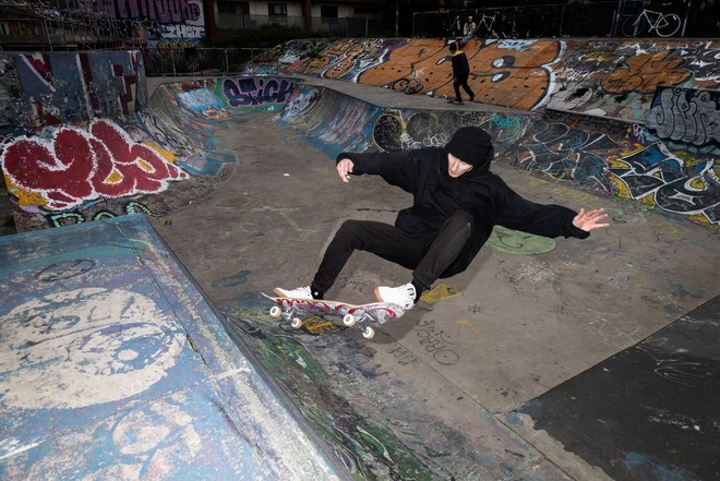 Мужчина в темной одежде и головном уборе выполняет трюк на скейтборде, отклоняясь под нужным углом в верхней части рампы.
