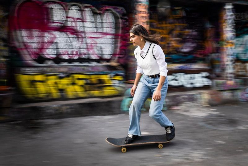 Девушка на скейтборде; покрытая граффити кирпичная стена на заднем плане размыта, в то время как девушка четко видна в кадре.