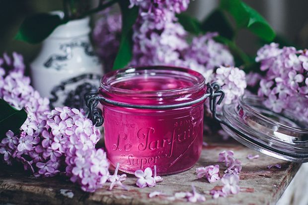 A jar of pink preserves on a wooden table surrounded by purple and white flowers. 