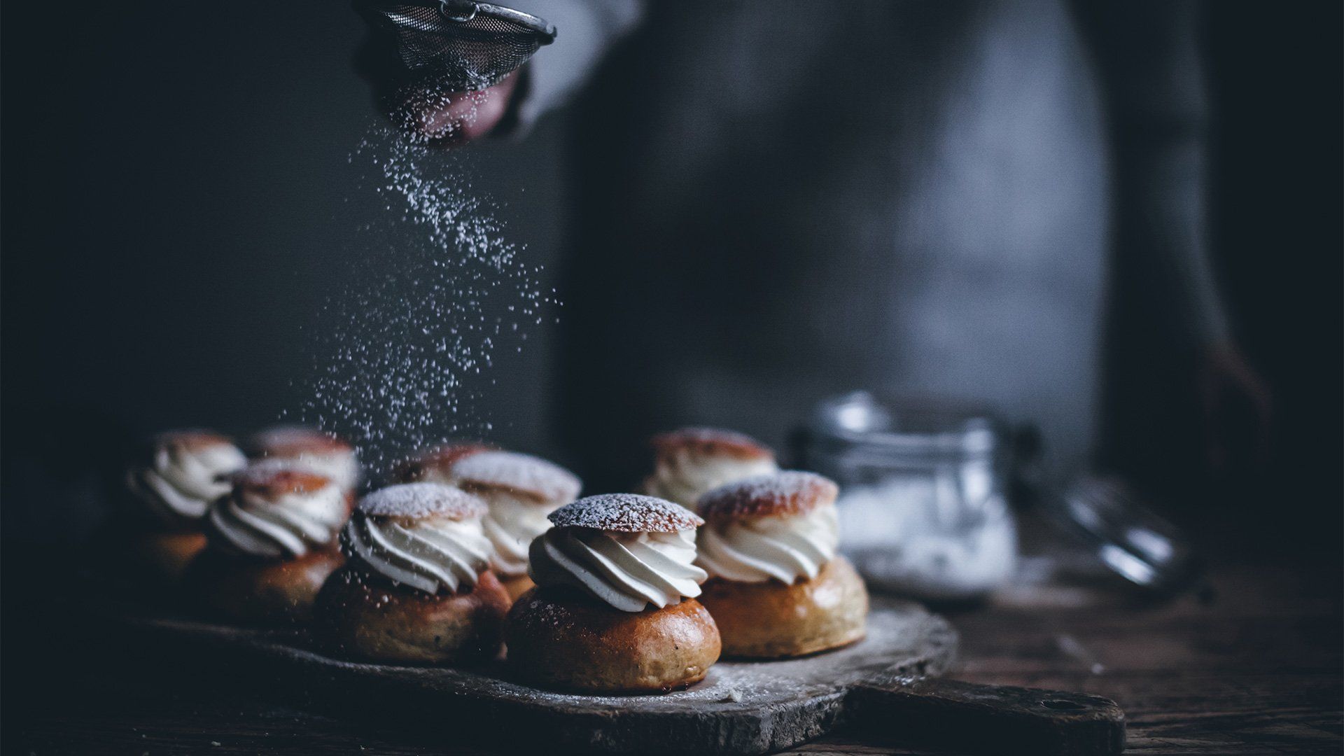 A person dusting icing sugar over pastries filled with piped cream. 