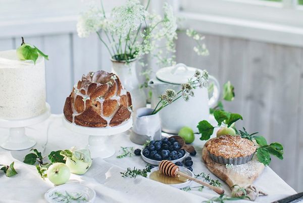 A table display on a white cloth, with cakes on stands, flowers in vases, and a honey dipper with a bowl of blueberries. 