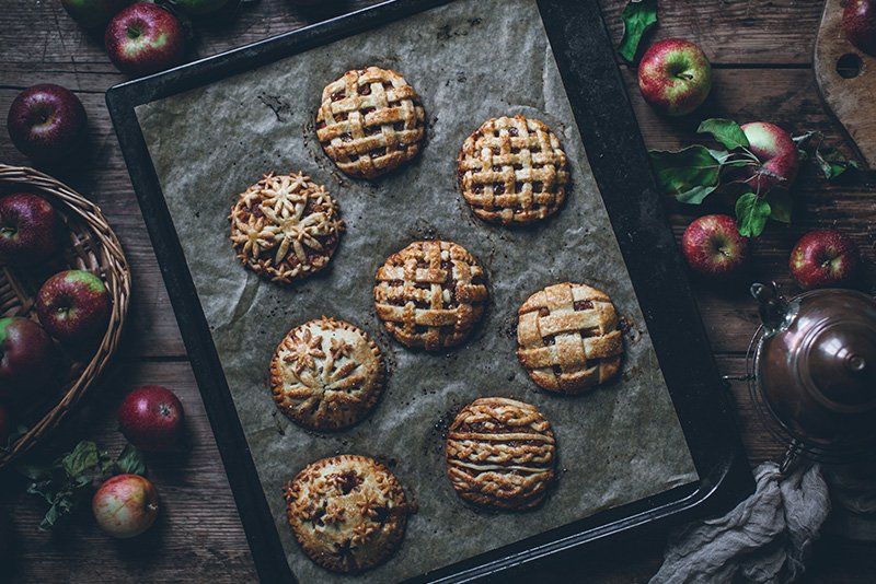 Eight small pastry topped pies on a tray surrounded by apples. 