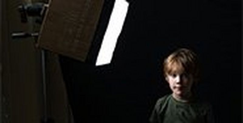 A boy stands looking at a photographer with a softbox made from a cardboard box to one side.