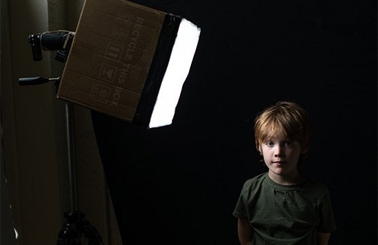 A boy stands looking at a photographer with a softbox made from a cardboard box to one side.