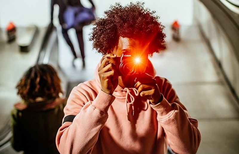 A man wearing a pink sweatshirt holds a Canon camera to his face. The flash casts an orange light.