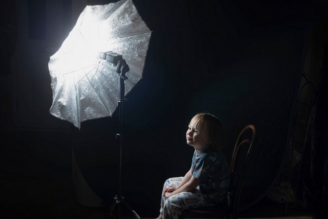 طفل جالس في الظلام مع وحدة فلاش ومظلة فضية تضيئان وجهه.