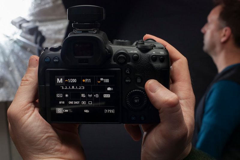 يُظهر الجزء الخلفي من كاميرا EOS R6 من Canon إعدادات درجة الإضاءة، مع رجل جالس أمام وحدة فلاش في الخلفية.