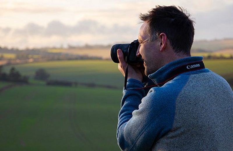 A landscape photographer stands under the branches of a tree in a frost-covered field bathed in sunshine.