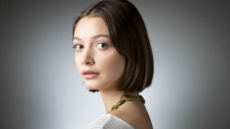 Portrait d'une femme portant un grand collier vert, sur un fond gris avec un éclairage spot blanc sur l'arrière-plan.