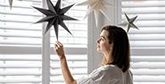A woman standing in front of shuttered windows touches a paper star hanging from the ceiling.