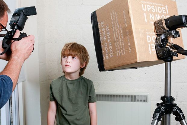 A boy stands looking at a photographer with a softbox made from a cardboard box to one side. 