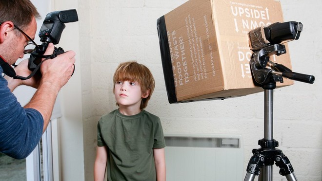 Мужчина фотографирует мальчика, стоящего перед вспышкой Speedlite, установленной на штативе за картонной коробкой.