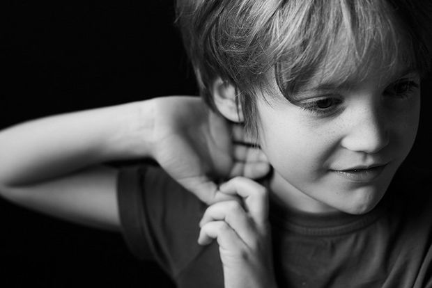  A black-and-white photo of a young boy raising one arm to his head, softly lit by a softbox.