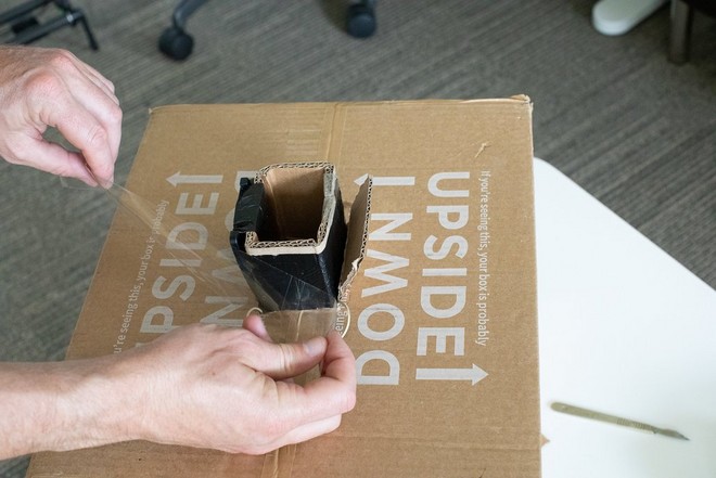 Мужчина с помощью скотча прикрепляет картонный конус к отверстию, вырезанному в нижней части большой картонной коробки.