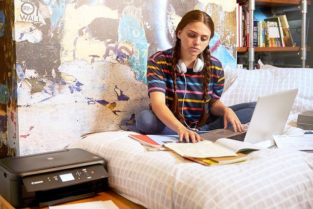 A teenage girl sits cross-legged on a bed, studying her textbooks. A Canon PIXMA TS5140 printer is on the table beside her.