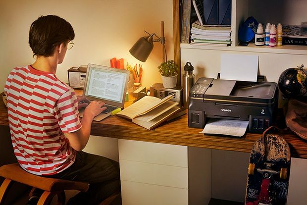 A teenage boy sits at his desk in his bedroom. A Canon G4500 printer is on the desk beside him.