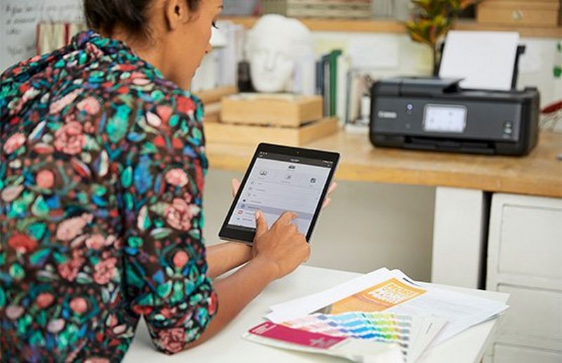 A woman with her back to the camera uses a tablet. A Canon printer is on a desk in the background.