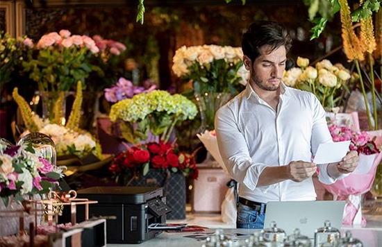 In a room filled with flowers, a man holds a print he has made using a Canon PIXMA G6040 that can be seen on the table in front of him.