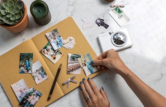 A woman's hands sticking wedding mini prints into a scrapbook, with a white Canon Zoemini S2 next to her.