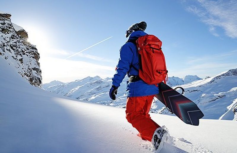 Um snowboarder atravessa a neve com a sua prancha de snowboard debaixo do braço, afastando-se da câmara. Fotografia de desportos de inverno de Richard Walch.