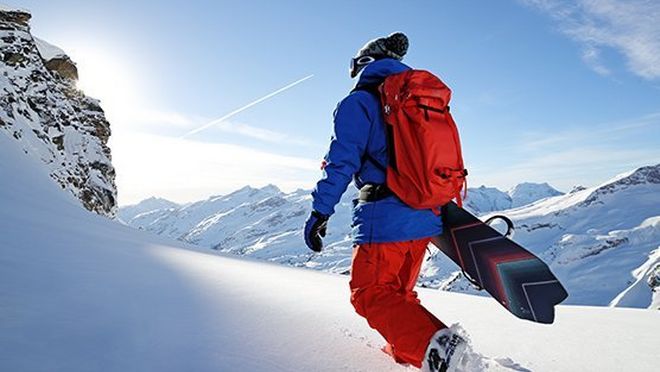 Ein Snowboardfahrer hält sein Snowboard unter dem Arm und läuft durch den Schnee, dabei bewegt er sich von der Kamera weg. Wintersportfoto aufgenommen von Richard Walch.