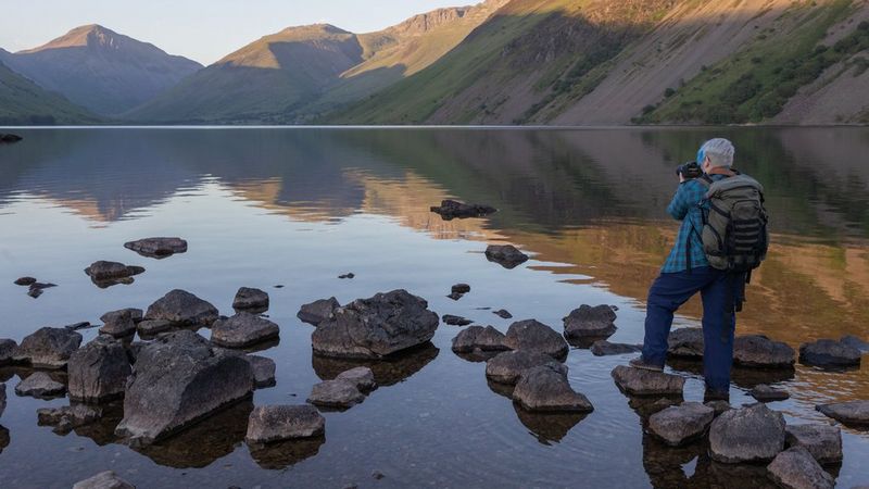 A female photographer stands on rocks in shallow water photographing a lake surrounded by mountains.