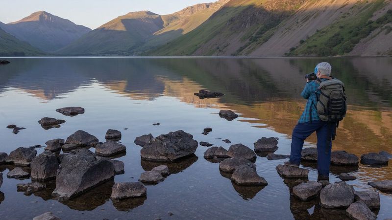 Женщина-фотограф стоит на камнях на мелководье и фотографирует озеро в окружении гор.