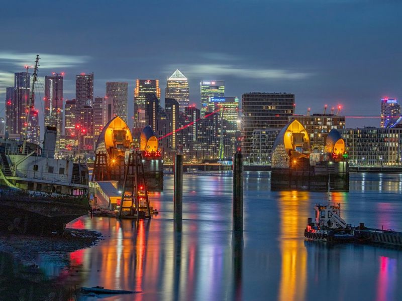 London's Thames Barrier at dusk, with the colourful lights of Canary Wharf visible in the background.