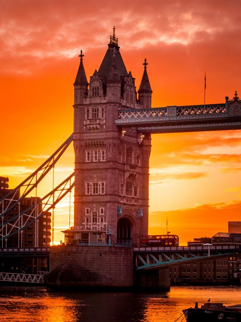 A double-decker bus crosses Tower Bridge in London, UK, at sunrise. The sky is orange in the background.