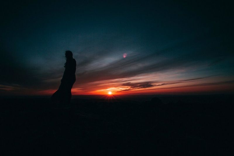 A figure stands silhouetted against a dramatic orange sunrise. 