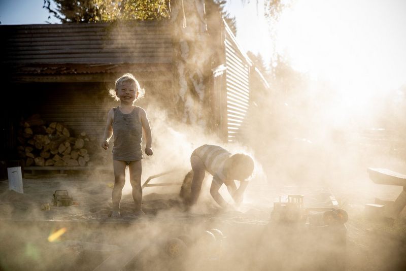 Two little boys playing in a sandpit in front of a log store, shrouded by dust.