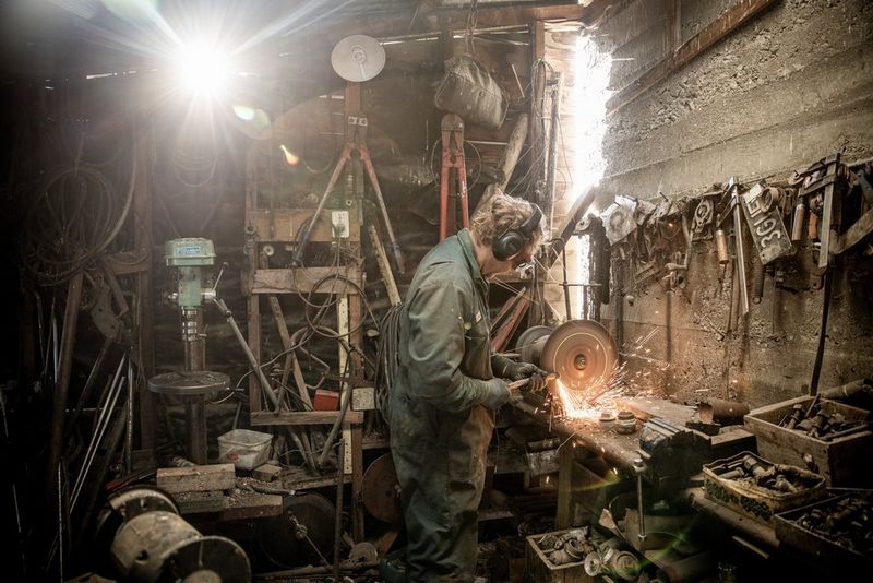 A man in a workshop using an angle grinder and surrounded by tools. The sun is shining through a gap in the back wall.