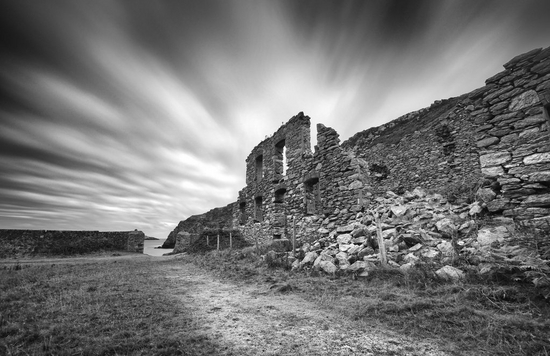 A black and white image of the ruins of a building, shot with a long exposure to blur the clouds in the sky.