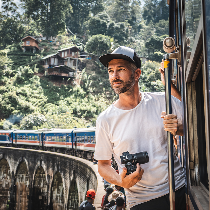 Photographer Martin Bissig leans out of a train that has stopped to pick up passengers on a viaduct in Sri Lanka. In his hand is a Canon EOS R10 and behind him a hill with small wooden houses built into it. Photographed by Monika Bissig on a Canon EOS R7. 
