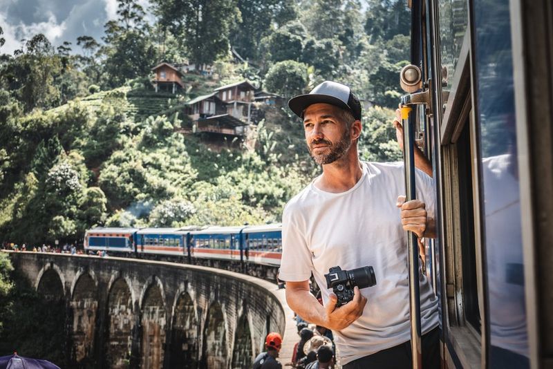Fotogrāfs Martins Bisigs (Martin Bissig) izliecas no vilciena, kas apstājies, lai uzņemtu pasažierus uz viadukta Šrilankā. Rokās viņam ir Canon EOS R10 kamera, un aiz viņa ir kalns, kurā iebūvētas mazas koka mājiņas. Uzņēmusi Monika Bisiga (Monika Bissig) ar Canon EOS R7. 