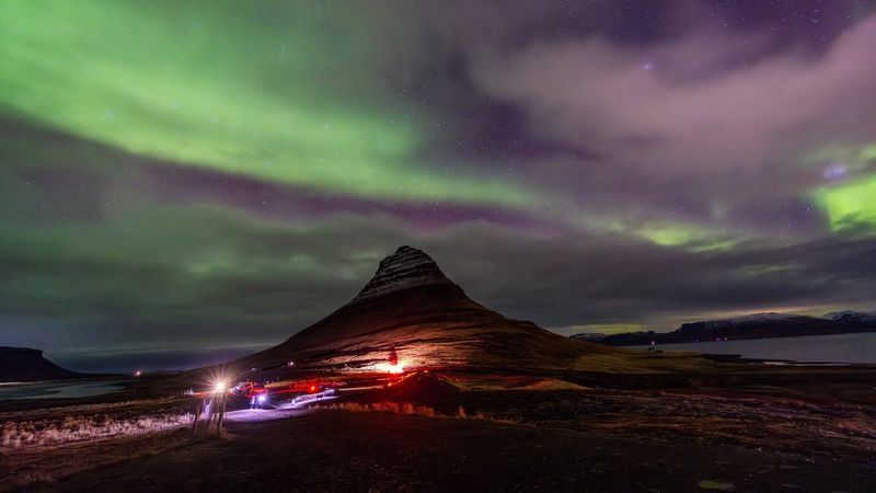 Zorza polarna w postaci zielonych smug na ciemnym nocnym niebie nad górą Kirkjufell w Islandii, z czerwonymi i fioletowymi światłami pojazdów i budynków widocznych u jej podnóża. Użyty sprzęt: aparat Canon EOS R5 z obiektywem Canon RF 14-35mm F4L IS USM. © Menna Hossam