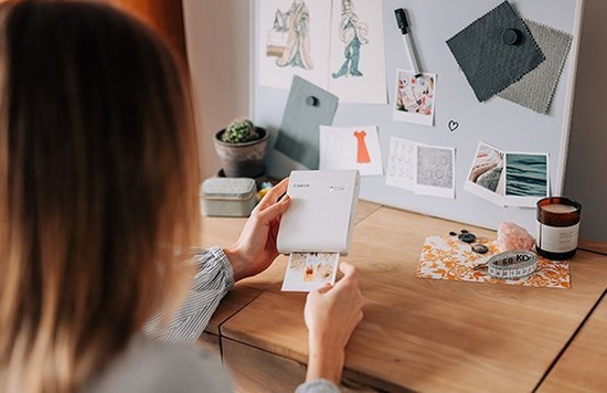 A woman prints out a picture on a Canon SELPHY Square QX10. Positioned upright on the desk in front of her is an outfit inspiration mood board.