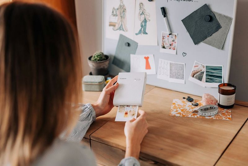 A woman prints out a picture on a Canon SELPHY Square QX10. Positioned upright on the desk in front of her is an outfit inspiration mood board. 