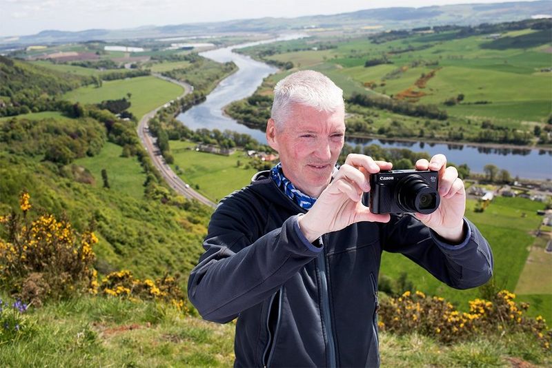 Alan Rowan stands on the side of a hill, with yellow gorse bushes, grassy fields and a river in the valley behind him. He holds a compact camera to take a photo of the natural view.