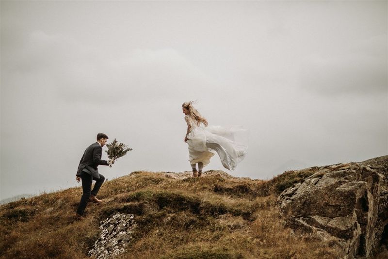A groom carries a bunch of flowers up a rugged hillside, his bride stands at the top, her dress flowing in the breeze.