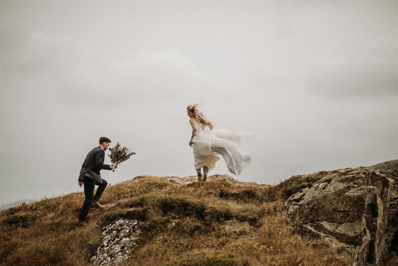 Жених с букетом поднимается по скалистому холму, невеста стоит на вершине, ее платье развевается на ветру.