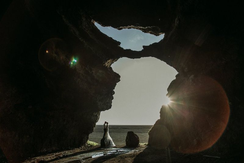 A newlywed couple embrace while standing on a small boulder. Shot from a distance, they are framed by a gap in a rocky outcrop.