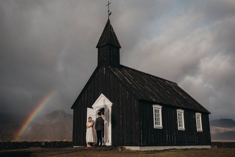 Молодожены стоят у белых дверей небольшой черной церкви. За ними на сером небе видно радугу.