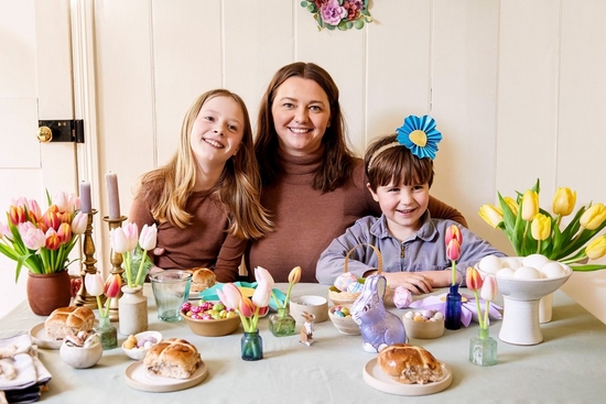 Crafter Hannah Bullivant sits between two children at a table decorated for spring with papercraft flowers, eggs and bunnies, vases of tulips and hot cross buns.