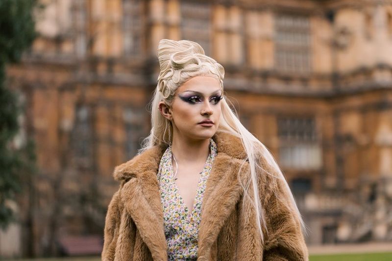  A portrait taken by Louis Painter of a drag queen, with long blonde hair and wearing a floral dress and brown fur coat, standing in front of an ornate building.